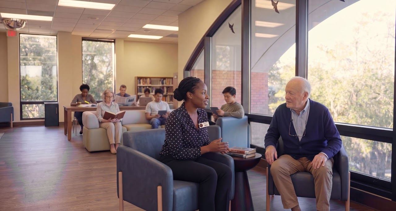 A library staff member and an older man sit talking in sturdy armchairs with armrests in a sunlit library seating area, with other patrons reading nearby.