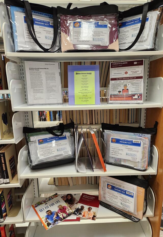 Library shelves displaying memory care kits in clear bags alongside informational brochures and pamphlets.