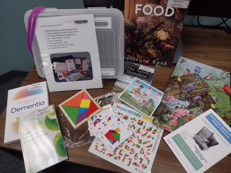 Contents of Memory Care Kit #1 spread on a table, including a tangram puzzle, jigsaw puzzle, picture books, music CDs, and dementia caregiver guides.